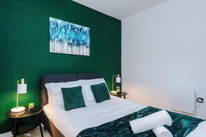 Une chambre moderne avec un mur vert, un lit avec une literie blanche et des coussins verts, un tableau au-dessus du lit et une lampe de chevet.