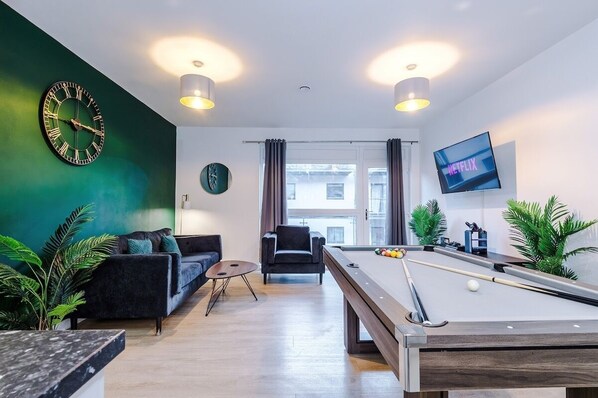 A modern living room with a pool table, a sofa, a chair, a clock, a television, and a view of the outside.