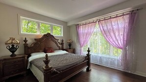 Une chambre à coucher avec un lit en bois, deux tables de chevet avec des lampes, une fenêtre donnant sur un paysage verdoyant et une autre fenêtre avec des rideaux violets.