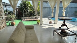 A modern living room with a beige sofa, a small table with books, and a view of a pool and garden. - Large house with floor (São Paulo)