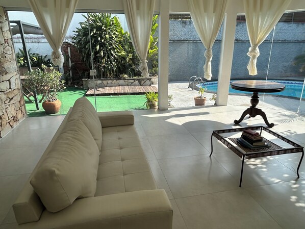 A modern living room with a beige sofa, a small table with books, and a view of a pool and garden.