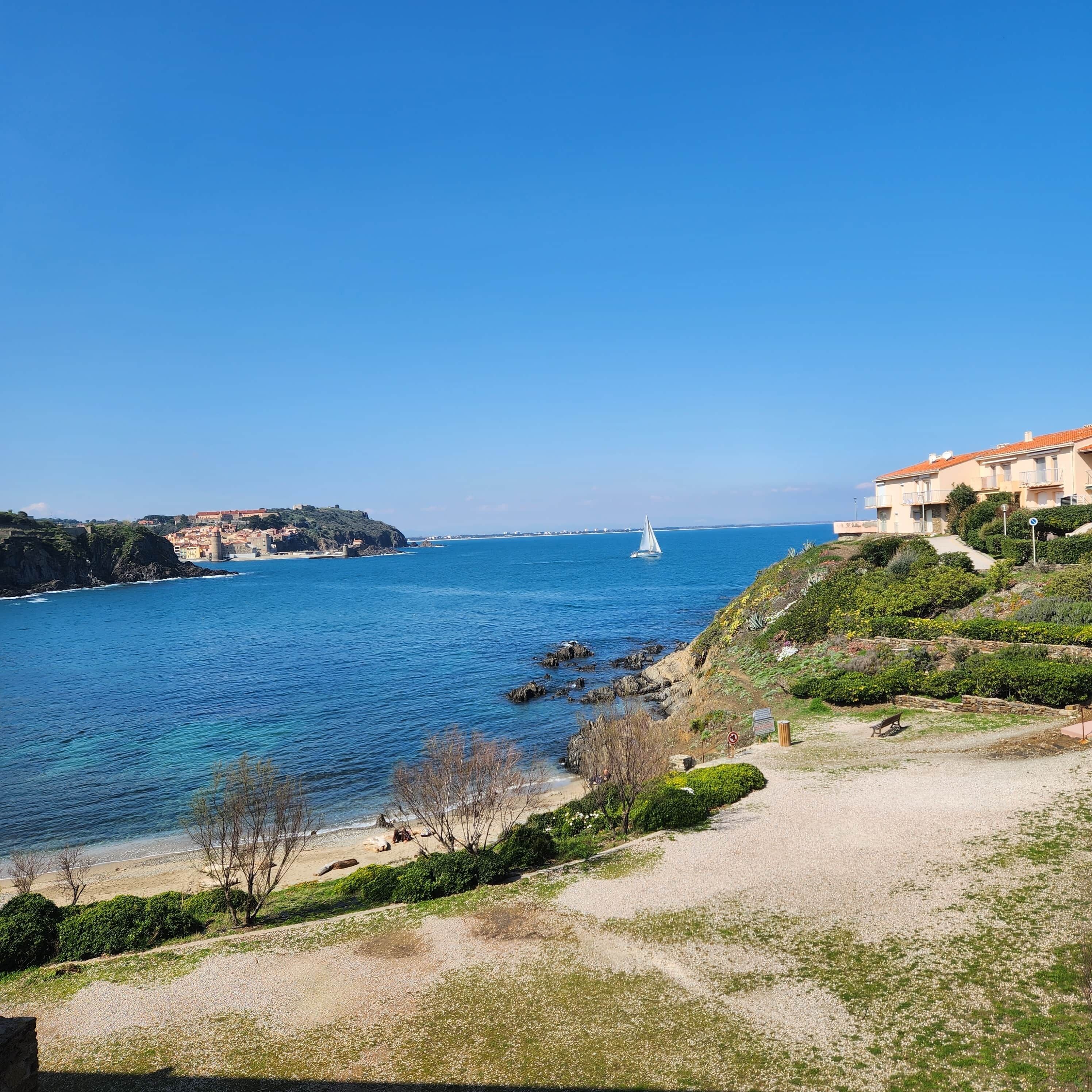 A coastal view with a clear blue sea, a sailboat, and a building with a red roof.