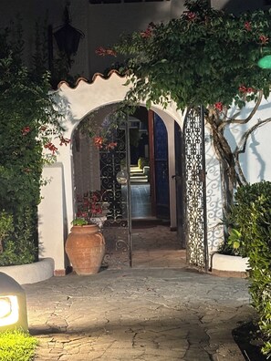 An arched entrance with a tiled pathway leading to a building, surrounded by greenery and a large pot.