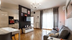 A modern living room with a grey sofa, a dining table, and a kitchen area with a TV and a clock. - Acasadigiò B&B - 400 meters from the Metro stop and 15 minutes from the center of Rome (Roma)