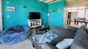 A living room with teal walls, a ceiling fan, a flat-screen TV on a white entertainment unit, a grey sofa, a blue bean bag, and a patterned rug. - Enjoy Passport to Paradise! (Christiansted)