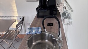 A kitchen with a stainless steel sink, a kettle, a microwave, and a coffee maker. There is a wall-mounted TV and a pegboard with books and a plant.