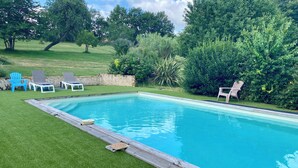 A rectangular swimming pool with a wooden deck edge, surrounded by a well-maintained lawn and various trees and shrubs.