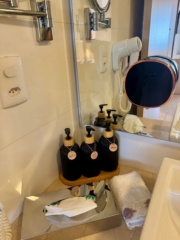 Baño con espejo, secador de pelo y tres botellas negras con tapones de madera etiquetadas como 'Personalize', 'Body Wash' y 'Shampoo'.
