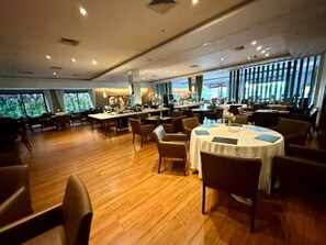 Un comedor amplio con múltiples mesas y sillas, ventanas grandes que ofrecen luz natural y vistas a la vegetación exterior.