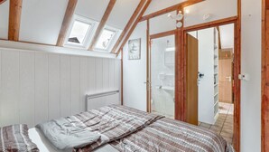 Ein Schlafzimmer mit schräg verlaufender Decke, Holzbalken, einem Bett mit gemusterten Textilien, einem Heizkörper und Blick ins Badezimmer durch eine Glastür.