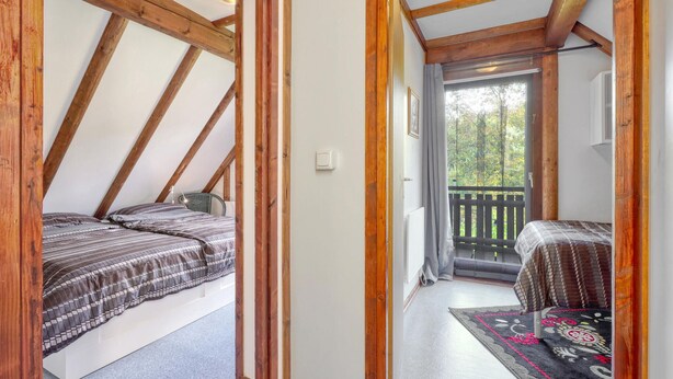 Ein Schlafzimmer mit geneigter Decke, einem Bett mit gemustertem Überwurf, einer Nachttischlampe und Blick durch eine Tür auf einen Balkon im Freien mit Geländer.