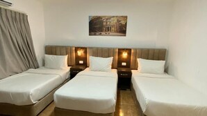 A hotel room with three single beds, a headboard with wall-mounted lamps, and a painting above the beds. - Petra Jewel Hotel (Wadi Musa)