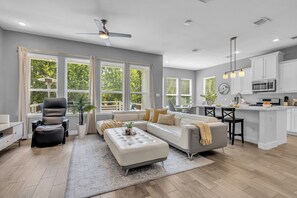 A modern living room with a sofa, ottoman, and a dining area with a kitchen in the background.