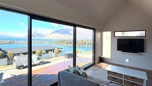 A modern living room with a large window, a grey sofa, a flat-screen TV mounted on the wall, and a wooden floor.
