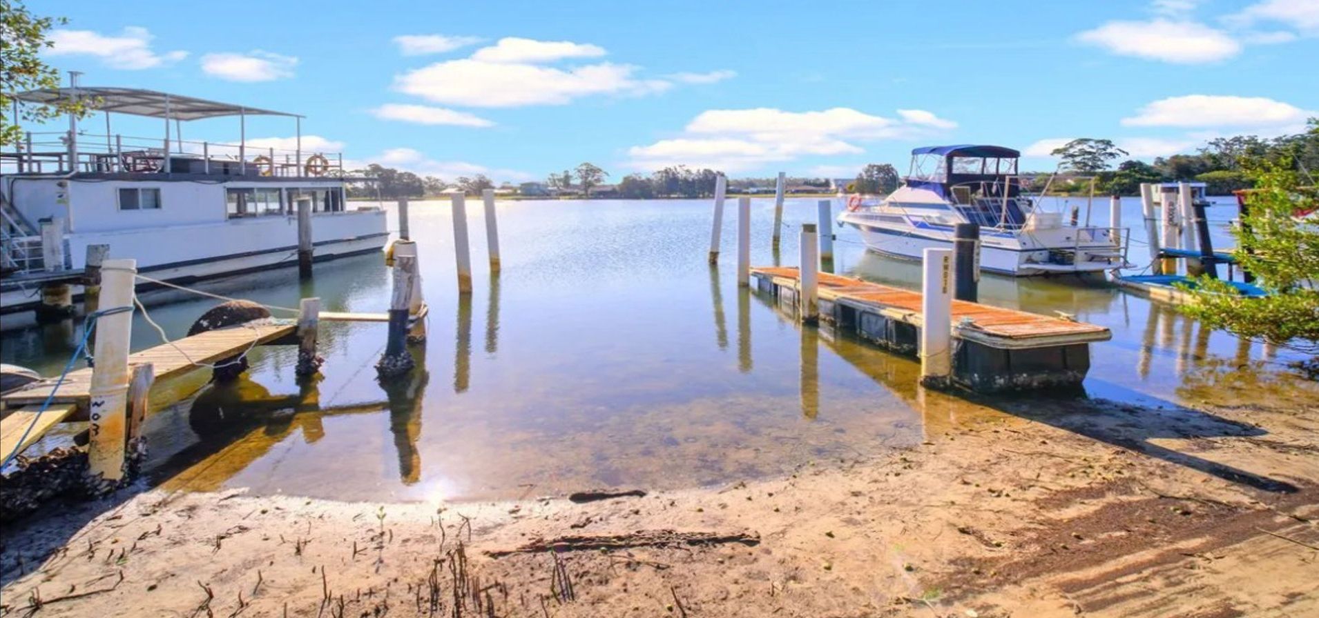 A calm waterfront with boats docked at wooden piers, a sandy shore, and a clear sky.