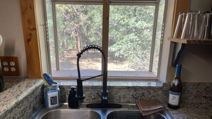 A kitchen with a double sink, a window with a view of trees, and a bottle of wine on the counter.