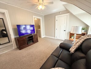 Un salon avec un téléviseur à écran plat posé sur un meuble en bois, un canapé en cuir noir, un ventilateur de plafond et une porte donnant sur une autre pièce.