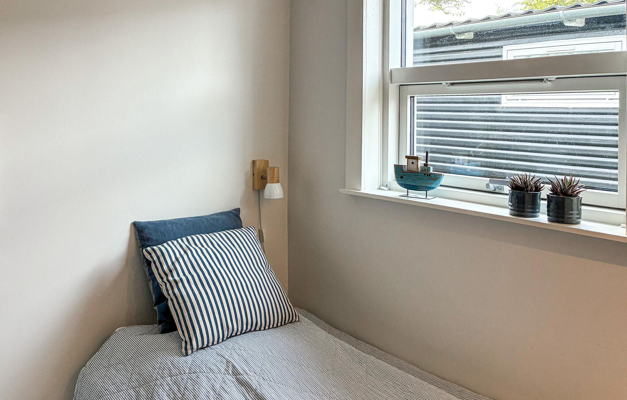 A corner of a room with a bed, a window with blinds, a wall-mounted lamp, and two potted plants on the windowsill.