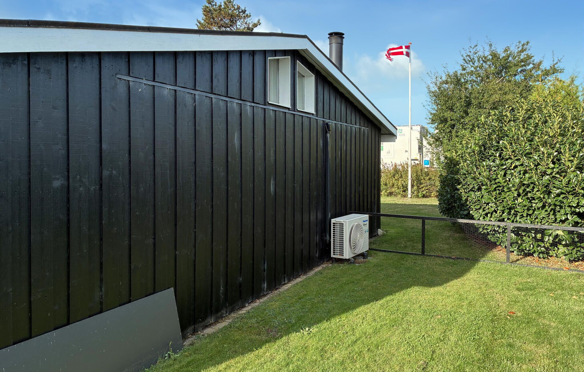 A black building with a chimney, a flag, and a hedge in the background.