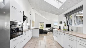 Une cuisine moderne avec des meubles blancs, un grand îlot central et un coin repas offrant une vue sur l’extérieur.