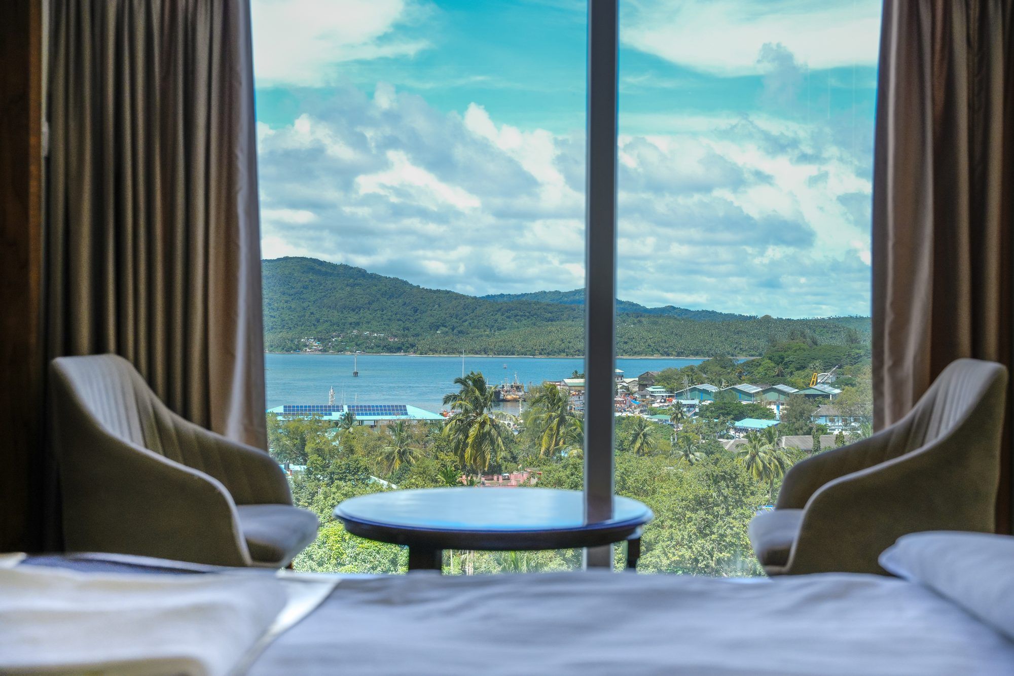 A hotel room with a bed, two armchairs, a small round table, and a view of a coastal landscape with mountains and a body of water.