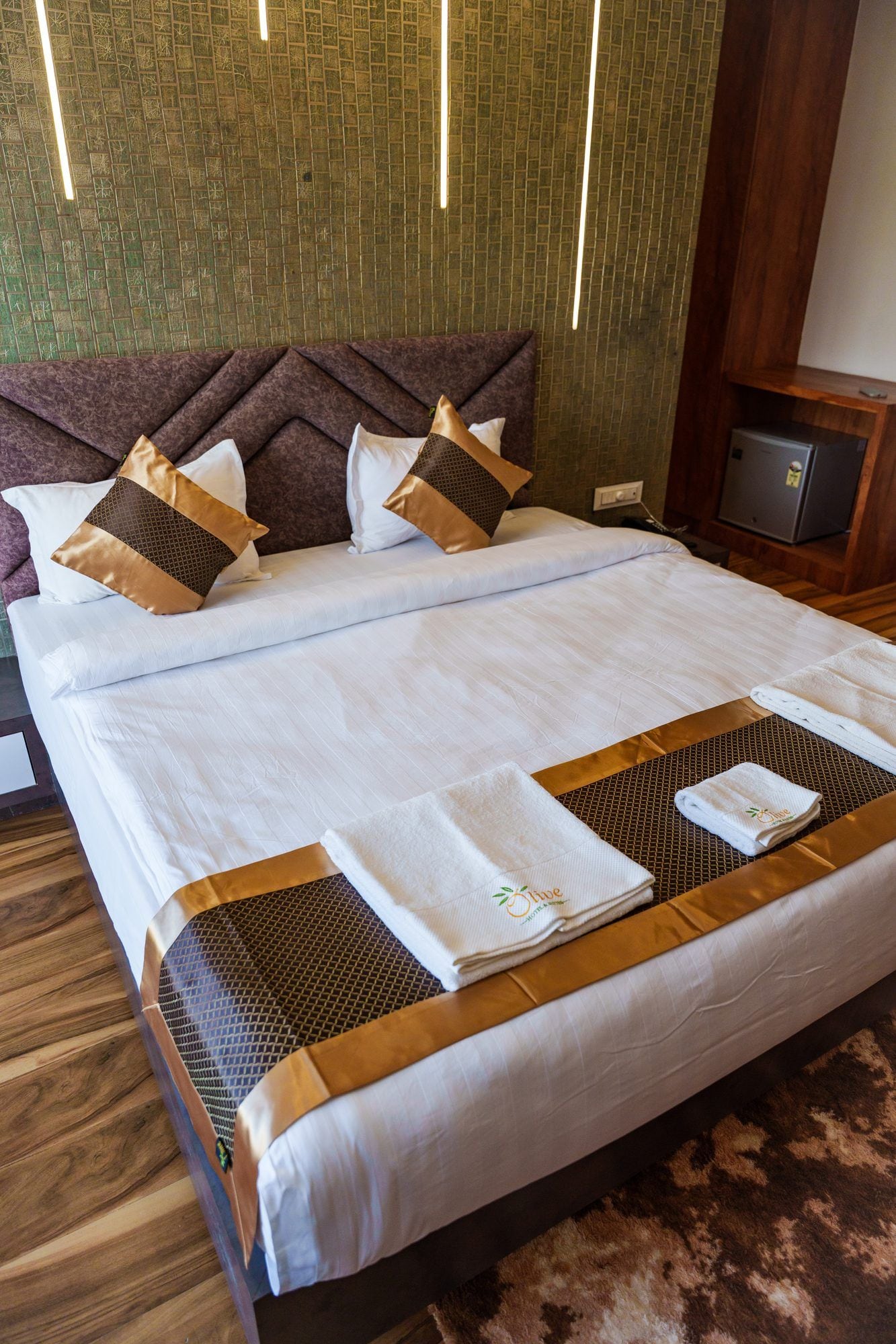 A double bed with a headboard, two pillows, a folded towel, and a small bedside table with a power outlet.