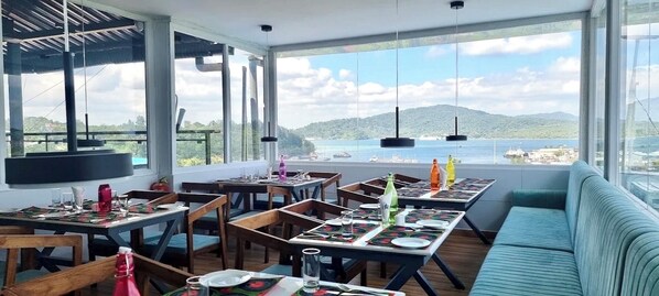 A dining area with tables set for four, large windows with a view of the sea and hills, and a blue sofa. - Olive Hotel & Suites (Port Blair)