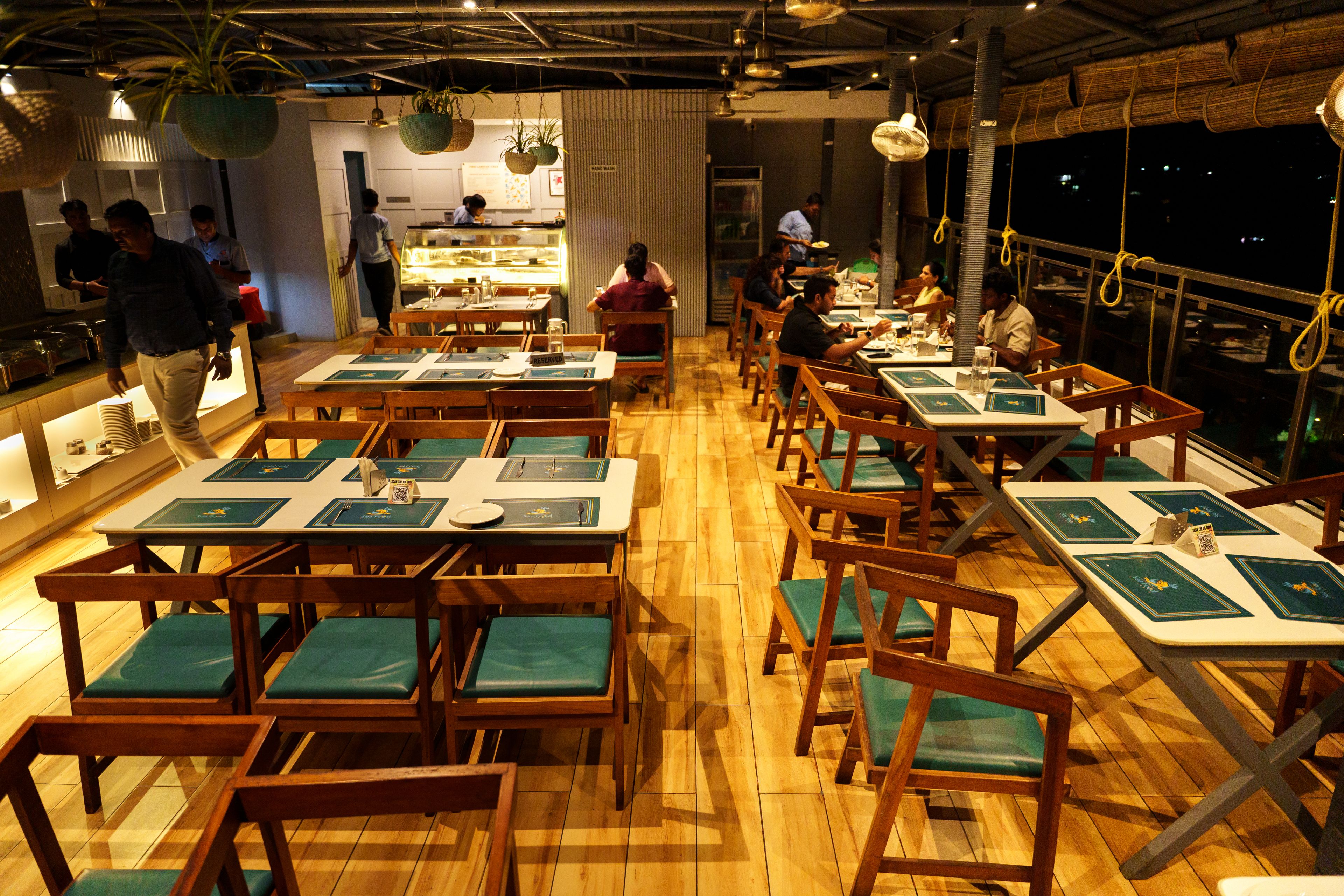 A restaurant with wooden tables and chairs, a kitchen area, and a view of the night sky.