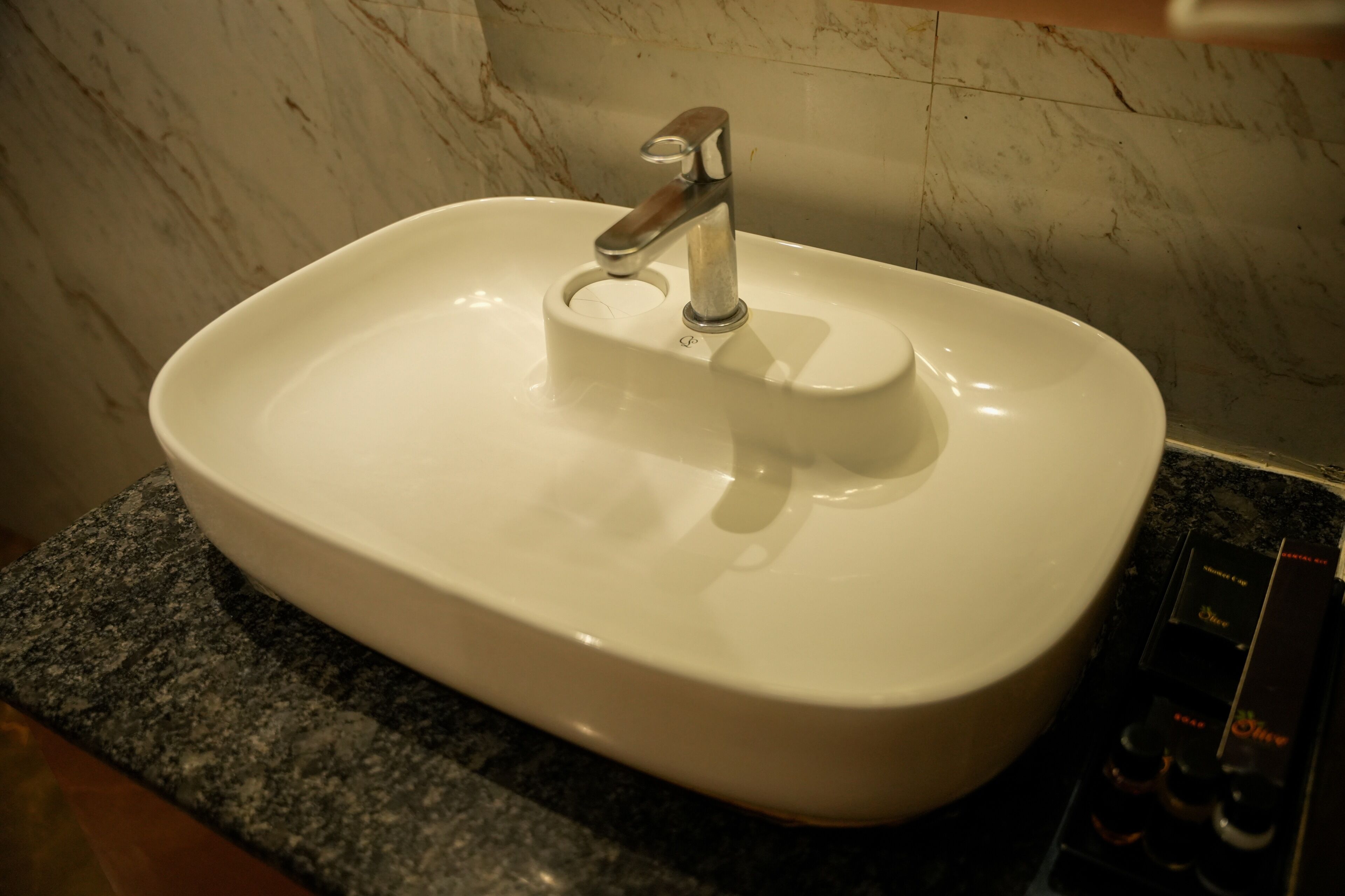 A white sink with a modern faucet, set on a granite countertop in a bathroom with marble walls.