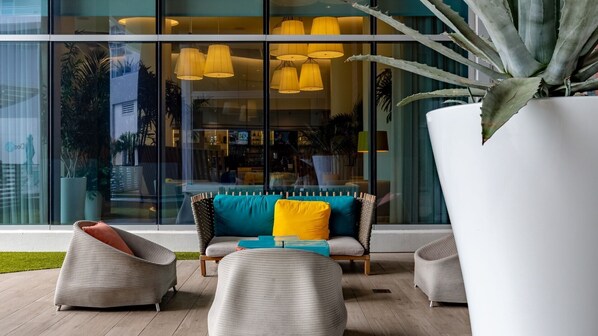 A modern outdoor seating area with wicker chairs and a sofa, a large potted plant, and a glass wall with interior lighting.