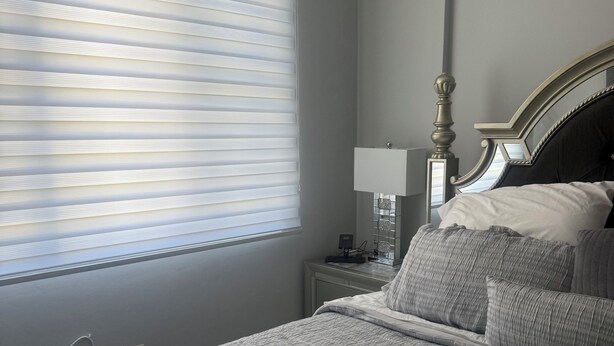Ein Schlafzimmer mit Bett, Nachttisch, Lampe und einem Fenster mit Jalousien.