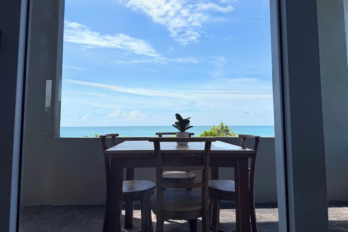 Une pièce dotée d’une grande fenêtre donnant sur une plage. On y trouve une table en bois, quatre chaises et un pot de fleurs posé sur le rebord de la fenêtre.
