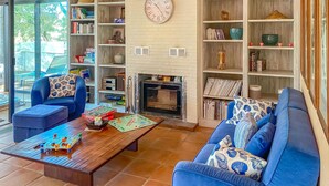 Una sala de estar con chimenea, sofás azules, una mesa de centro con juegos de mesa y una estantería llena de libros y adornos.