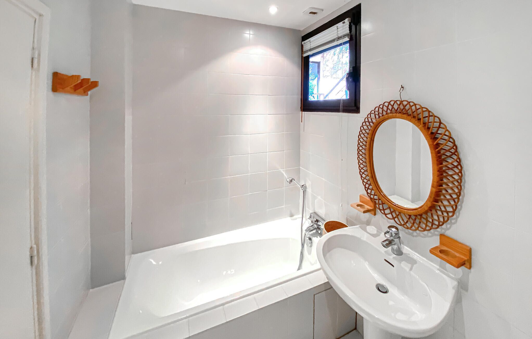 A modern bathroom with a white bathtub, a sink with a wooden frame mirror, and a window with a view of greenery.