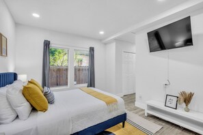 A bedroom with a bed, a TV mounted on the wall, a window with curtains, and a door. - Modern 4BD Retreat- 3 Ensuite BRs - Gated Parking - Walk to Metro (Houston)