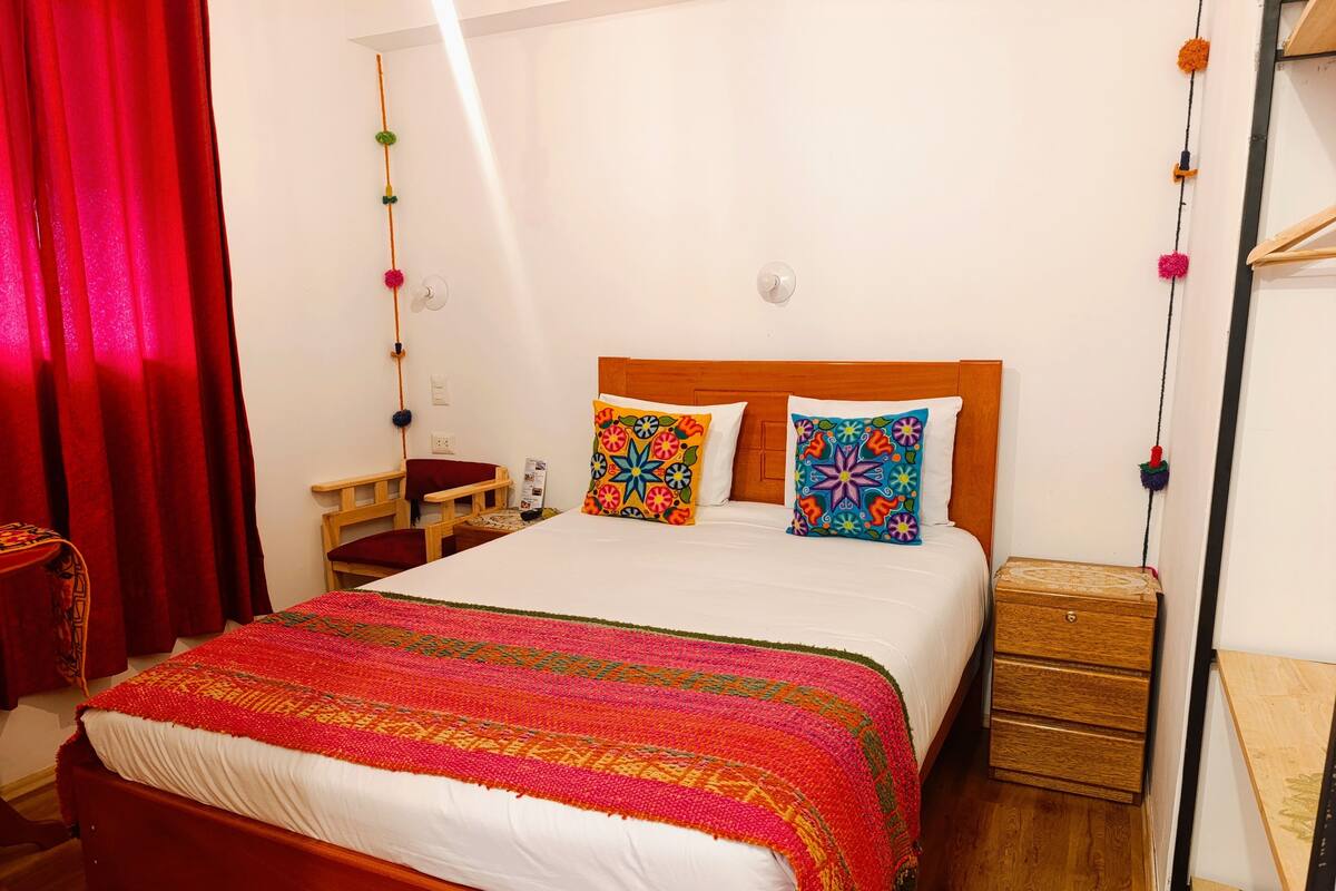 A neatly made bed with colorful pillows, a wooden nightstand, and a red curtain.