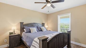 A bedroom with a bed, two bedside tables, a ceiling fan, a window with blinds, and a view of greenery outside.