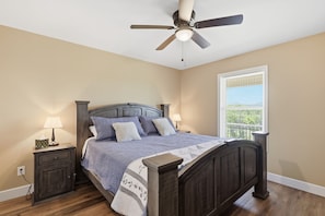 A bedroom with a bed, two bedside tables, a ceiling fan, a window with blinds, and a view of greenery outside.