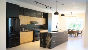 A modern kitchen with dark cabinetry, a marble island, and a dining area with a large window. - New Luxury Palm Springs Home for Family & Friends (Palm Springs)