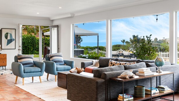 A modern living room with a large window, a grey sofa, two armchairs, and a coffee table with books and a globe.