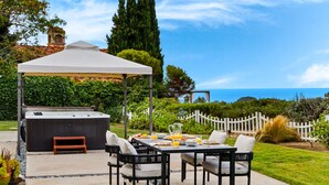 An outdoor patio with a grill, dining table, and chairs, surrounded by greenery and a white fence.