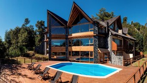 Ein modernes mehrgeschossiges Haus mit großem Schwimmbecken, Holzterrasse und einer Bergkulisse.