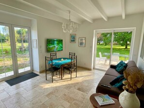 A spacious living room with a dining area, a sofa, a TV, and a view of a garden through glass doors. - Domaine des Anes - Gîte des Mûriers (Tugéras-Saint-Maurice)