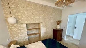 Un dormitorio con pared de piedra, cama con ropa de cama blanca y azul oscuro, cómoda de madera y lámpara colgante.