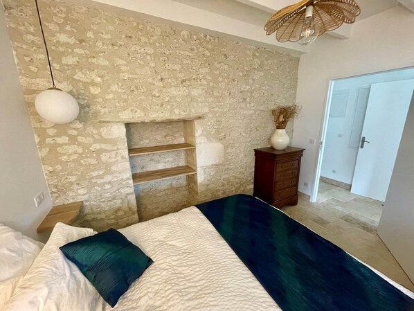 A bedroom with a stone wall, a bed with white and dark blue bedding, a wooden dresser, and a hanging light fixture.