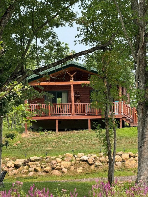 A wooden cabin with a porch, surrounded by trees and a stone wall.