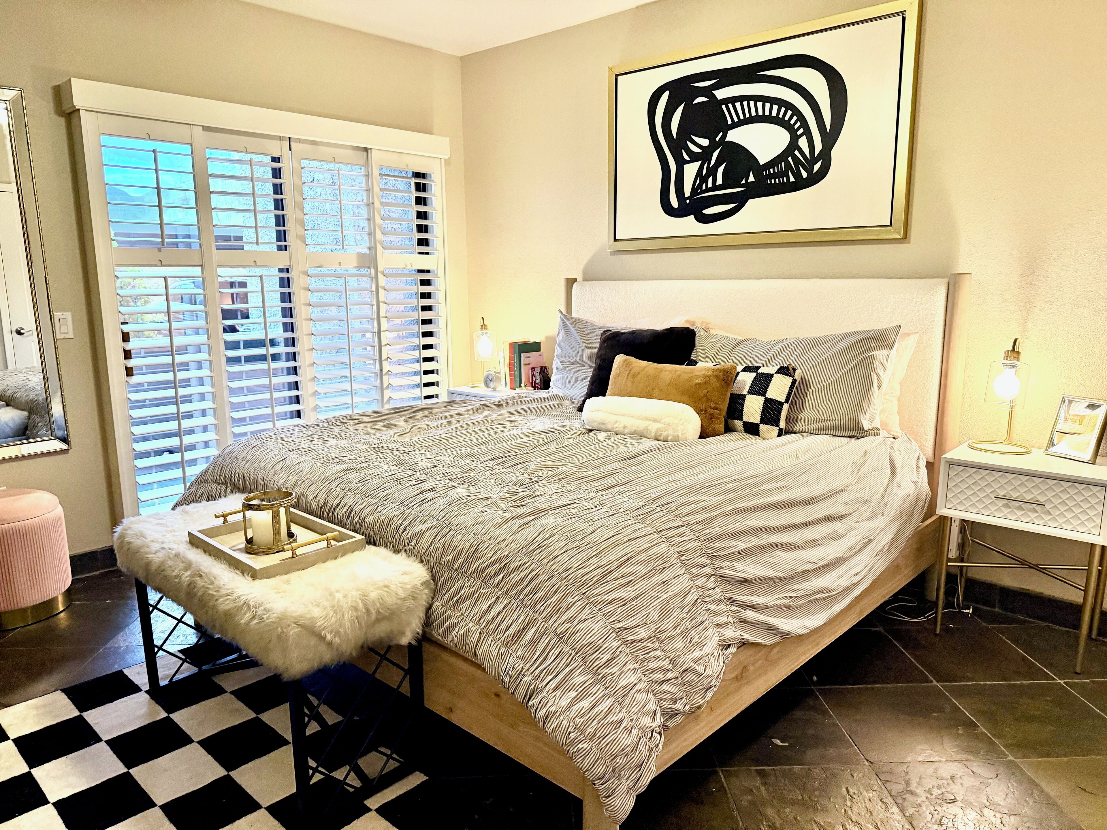 A bedroom with a large bed, a bedside table with a lamp, a framed picture, and a window with blinds.