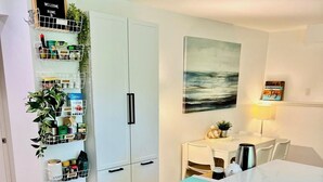 Eine moderne Küche mit weißen Schränken, einem eingebauten Geschirrspüler und Holzboden. Eine Wandablage zeigt verschiedene Küchenutensilien und ein Gemälde vom Meer.
