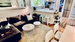 Ein modernes Wohnzimmer mit einer Couch, einem Couchtisch und Sesseln. Es gibt eine Küchenzeile mit Kühlschrank, Mikrowelle und Spüle.