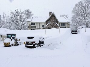 Zona residencial cubierta de nieve, con una casa, vehículos estacionados y un quitanieves.
